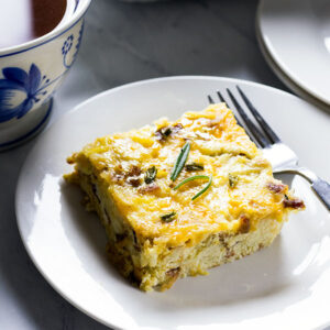 Slice of egg, potato and cheese casserole garnished with rosemary leaves on a white plate, with a fork and a blue and white mug of coffee