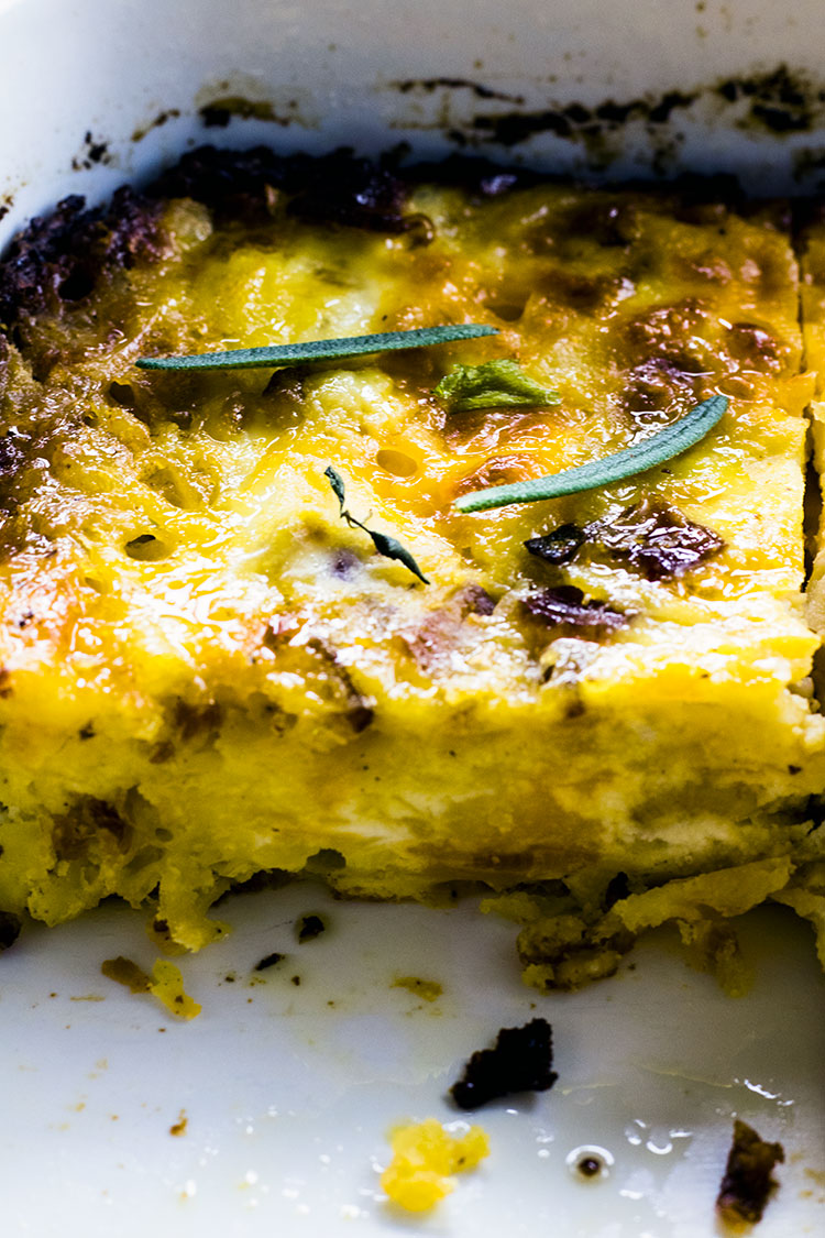 Close up of egg, potato and cheese breakfast casserole garnished with rosemary leaves sitting in a white casserole dish with slice taken out