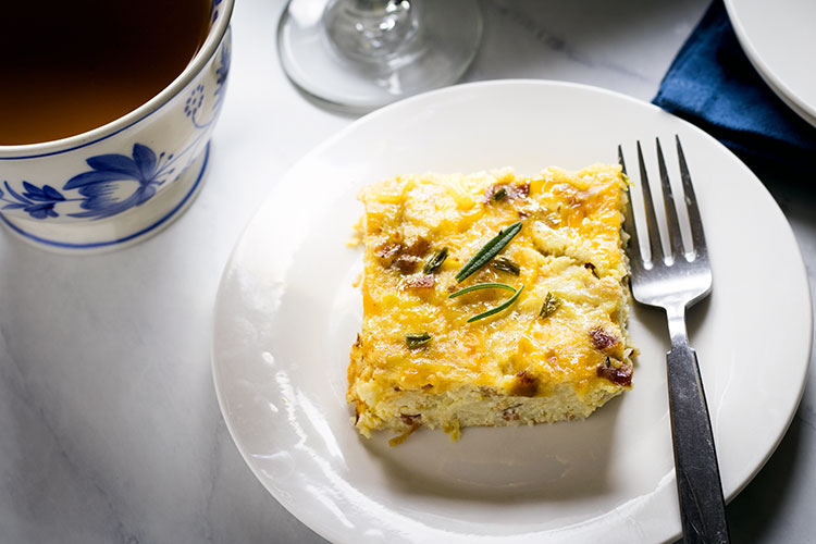 Slice of egg, potato and cheese casserole garnished with rosemary leaves on a white plate, with a fork and a blue and white mug of coffee