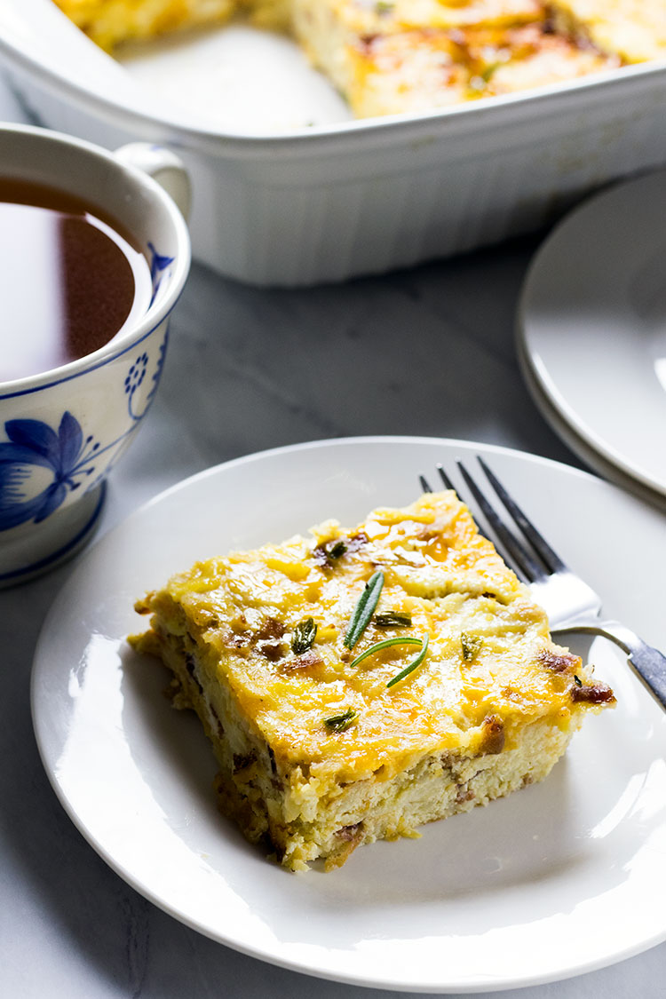 Slice of egg, potato and cheese casserole garnished with rosemary leaves on a white plate, with a fork and a blue and white mug of coffee