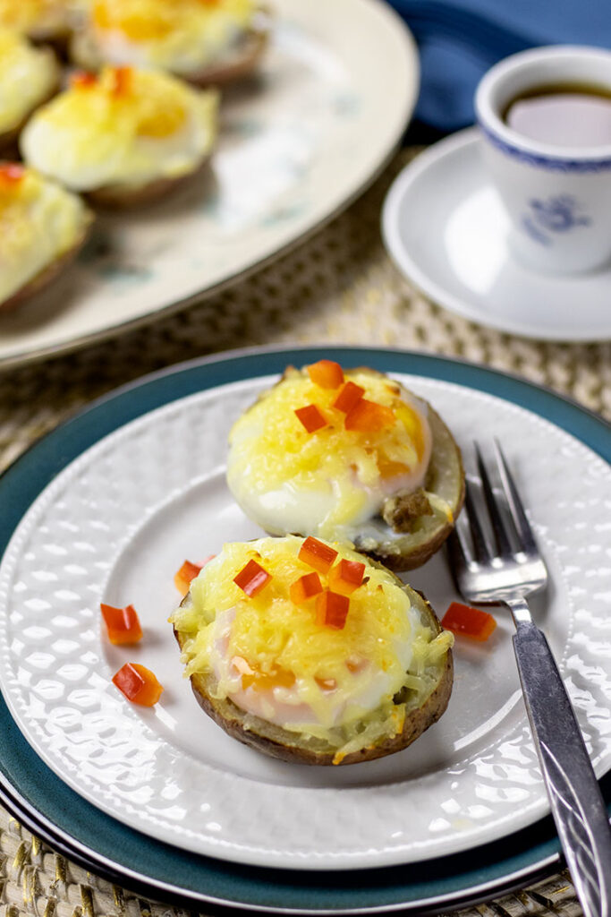 Two potato skins with turkey sausage, poached eggs, melted cheese and chopped red pepper on a white plate sitting on top of a turquoise plate with a fork. A blue napkin, cup of coffee and platter of potato skins are sitting in the background.