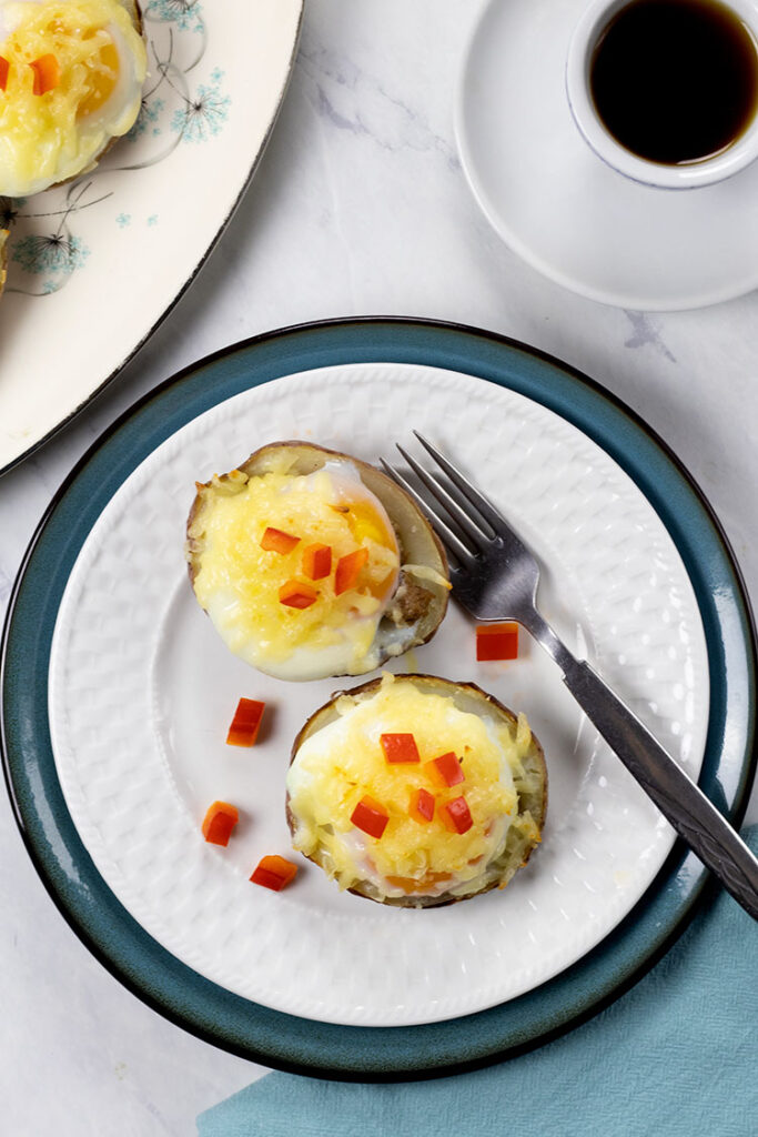 Overhead photo of two potato skins with turkey sausage, poached eggs, melted cheese and chopped red pepper on a white plate sitting on top of a turquoise plate with a fork. A blue napkin, cup of coffee and platter of potato skins are also in the photo.
