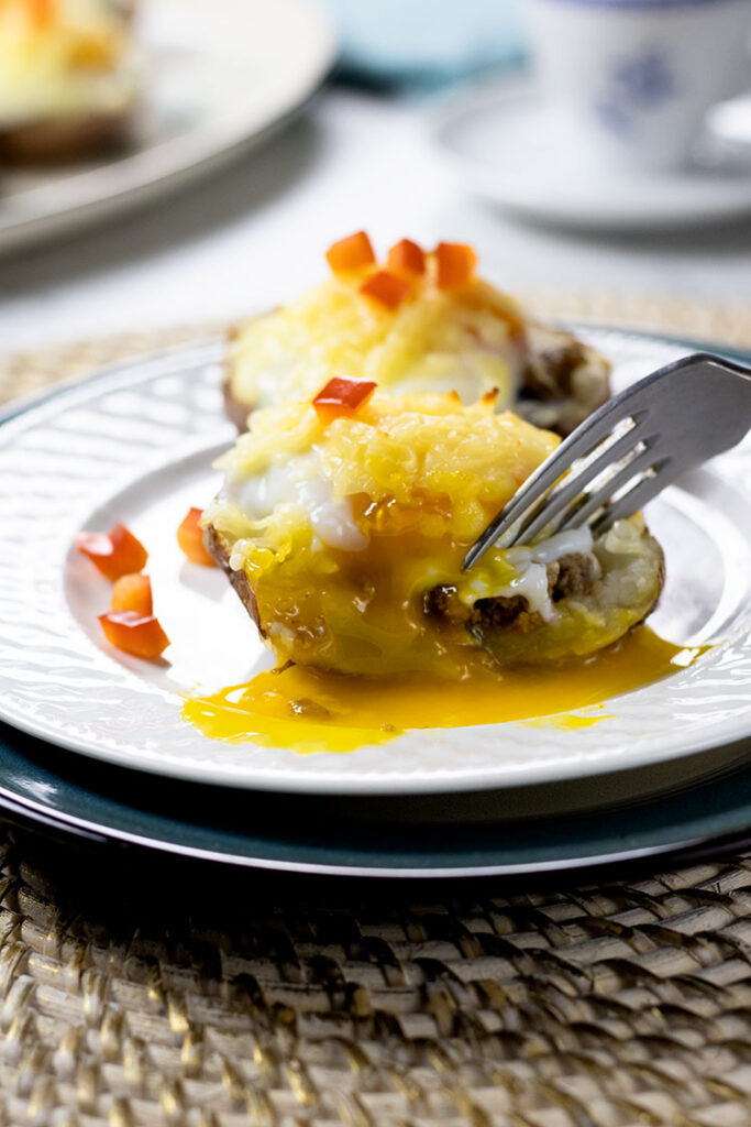 Two potato skins with turkey sausage, poached eggs, melted cheese and chopped red pepper on a white plate sitting on top of a turquoise plate with a fork cutting into one of the potato skins and egg yolk is running out. A cup of coffee and platter of potato skins are sitting in the background.