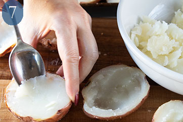 Hand scooping the flesh out of half of a potato. Hollowed out potato halves and a white bowl containing potato flesh are also shown.