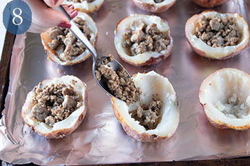 Cookie sheet containing hollowed out potato skins containing cooked turkey sausage. Turkey sausage is being spooned into one potato skin.