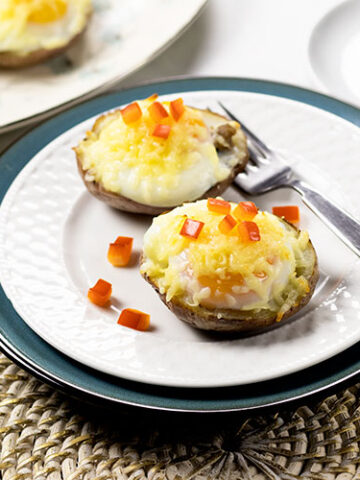 Two potato skins with turkey sausage, poached eggs, melted cheese and chopped red pepper on a white plate sitting on top of a turquoise plate with a fork. A cup of coffee and platter of potato skins are sitting in the background. A turquoise napkin sits in the foreground.