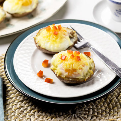 Two potato skins with turkey sausage, poached eggs, melted cheese and chopped red pepper on a white plate sitting on top of a turquoise plate with a fork. A cup of coffee and platter of potato skins are sitting in the background. A turquoise napkin sits in the foreground.