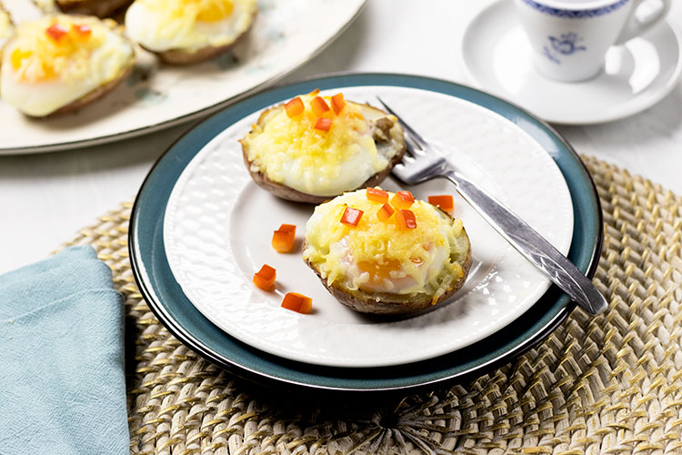 Two potato skins with turkey sausage, poached eggs, melted cheese and chopped red pepper on a white plate sitting on top of a turquoise plate with a fork. A cup of coffee and platter of potato skins are sitting in the background. A turquoise napkin sits in the foreground.