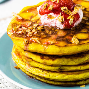 Stack of orange pancakes sitting on a turquoise plate topped with yogurt, fresh strawberries and homemade granola..