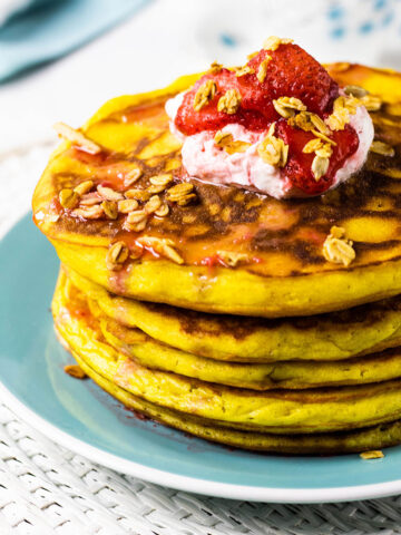 Stack of orange pancakes sitting on a turquoise plate topped with yogurt, fresh strawberries and homemade granola..