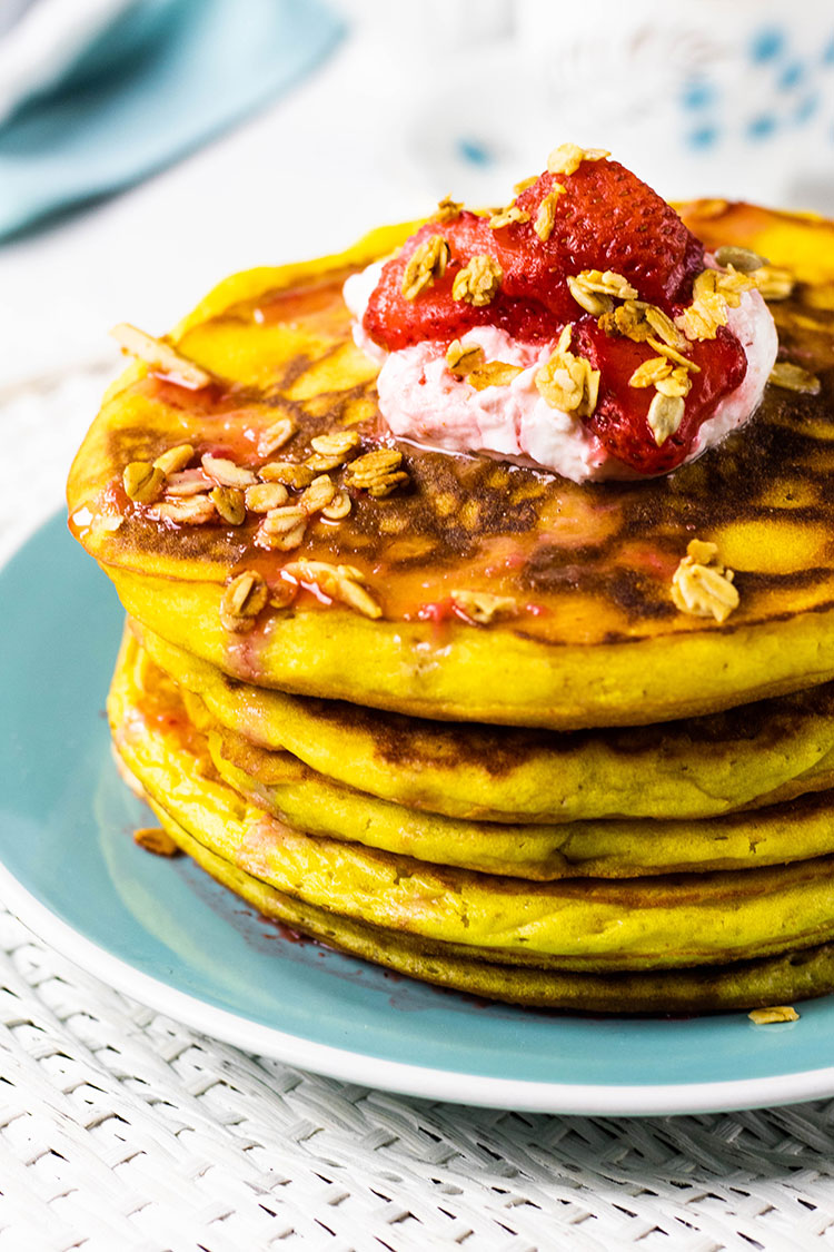 Stack of orange pancakes sitting on a turquoise plate topped with yogurt, fresh strawberries and homemade granola..