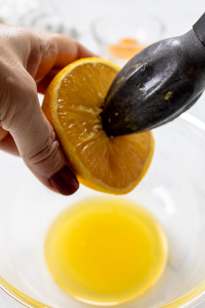 A cook's hand is holding an orange half and squeezing it with a metal citrus squeezer over a glass bowl.