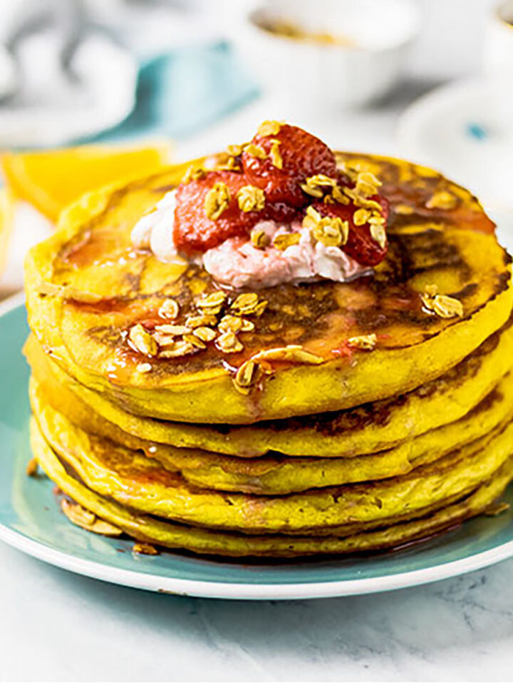 Stack of orange pancakes sitting on a turquoise plate topped with yogurt, fresh strawberries and homemade granola. Fresh oranges, a turquoise and white napkin and cup of coffee or tea are in the background.