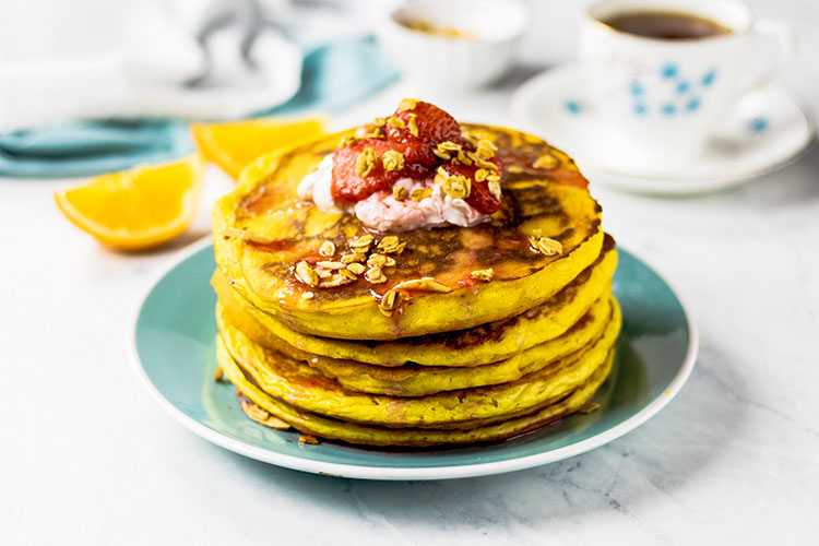 Stack of orange pancakes sitting on a turquoise plate topped with yogurt, fresh strawberries and homemade granola. Fresh oranges, a turquoise and white napkin and cup of coffee or tea are in the background.