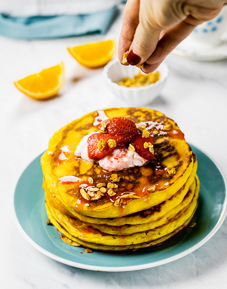 Stack of orange pancakes sitting on a turquoise plate topped with yogurt and fresh strawberries. The cook's hand is coming down from the top and sprinkling homemade granola over the pancakes. Fresh oranges and a turquoise napkin are in the background.