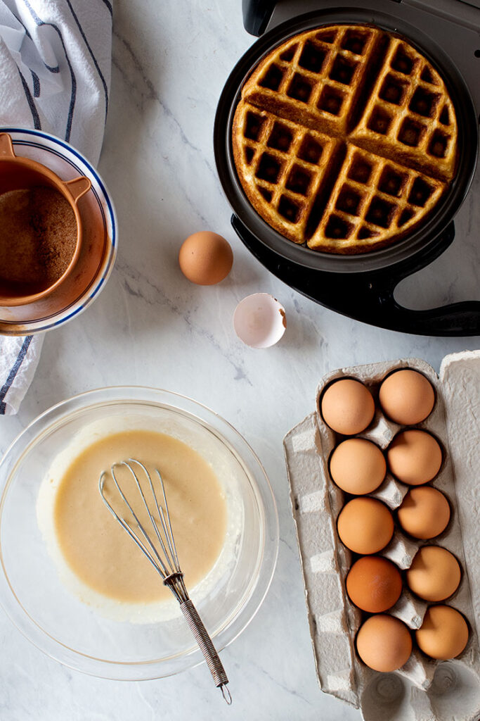 Overhead photo of a carton of eggs, waffle batter, eggshells, and waffles in a waffle iron