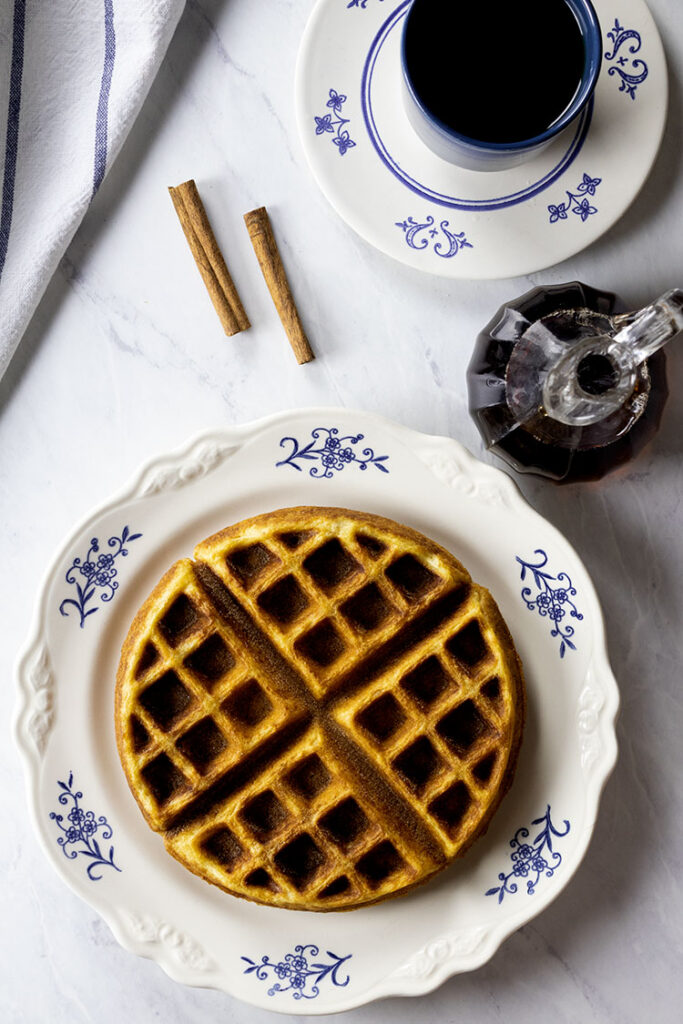 Overhead photo of a buttermilk waffle sitting on a white plate with blue flowers. A small pitcher of hot syrup, coffee cinnamon sticks and a blue and white towel are also in the photo.