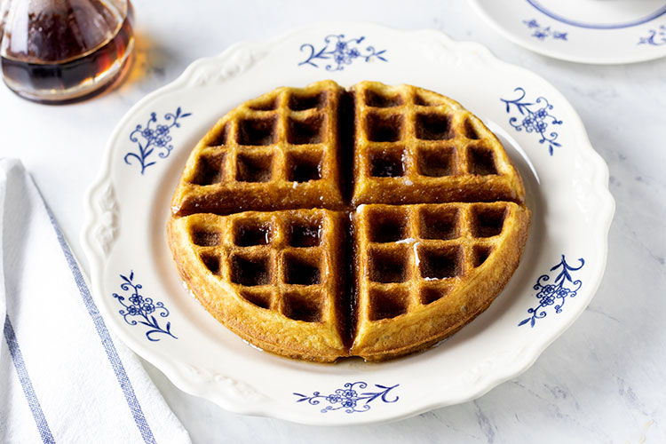 Horizontal photo of a buttermilk waffle with syrup on it sitting on a white plate with blue flowers, a folded blue and white napkin sits on the left side of the plate. A flask of syrup sits in the background.