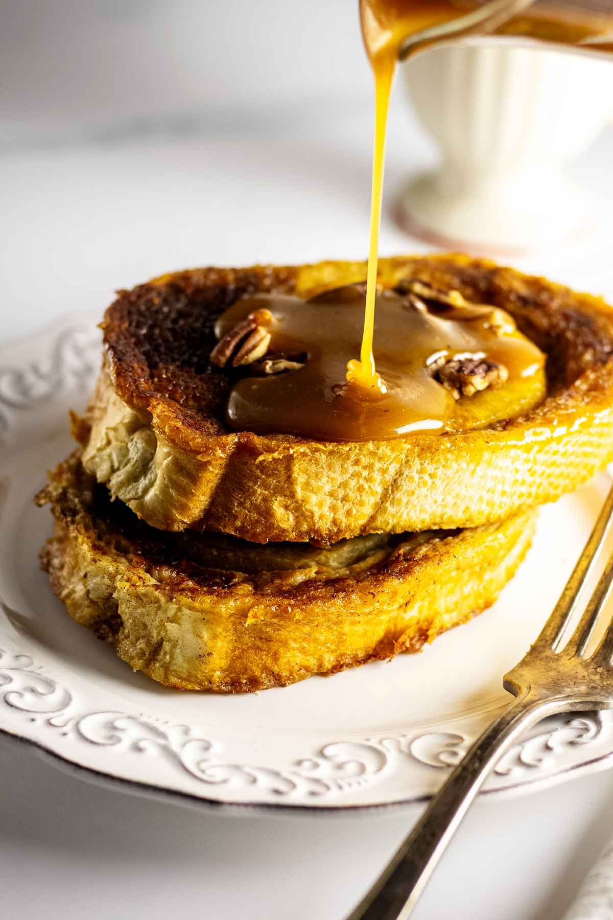 Caramel apple French toast topped with apple rings and chopped pecans on a white plate with fork. Caramel syrup is being poured over the top.