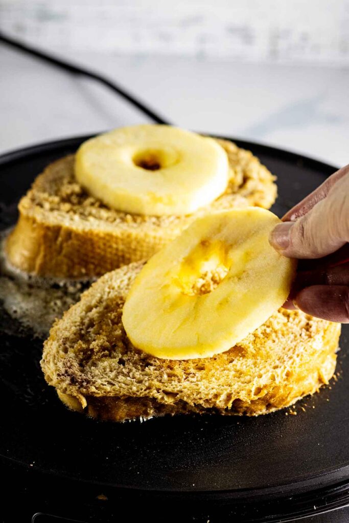 Two slices of French toast cooking on a griddle. A sliced apple ring is being placed on top of one of the slices.