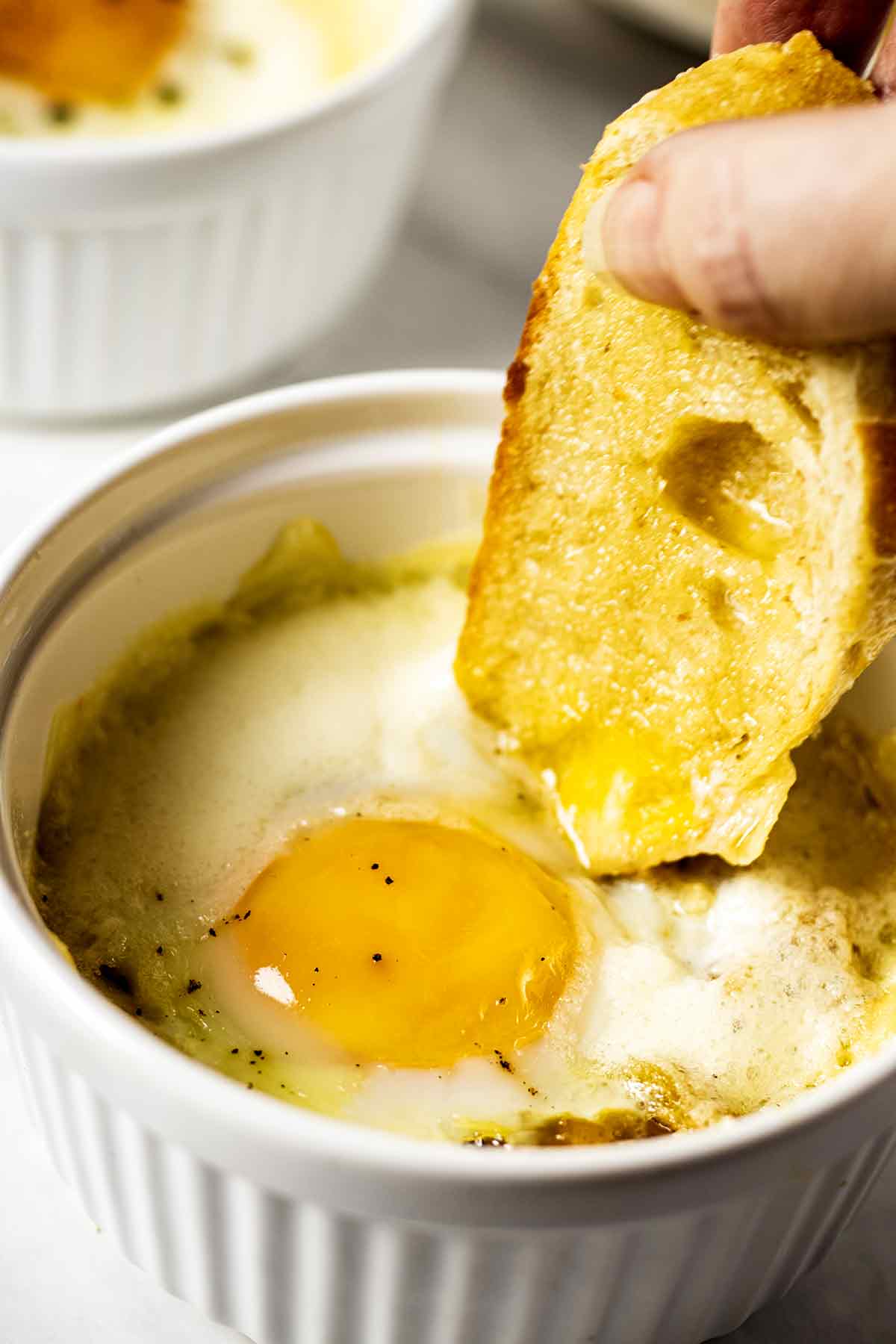 Toast being dipped into Swiss baked egg in a white ramekin