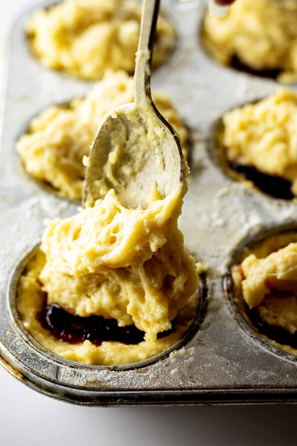 Batter being spooned over fruit preserves in a muffin tin