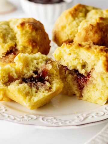 Three biscuit muffins on a white plate with one cut in half exposing the jam inside