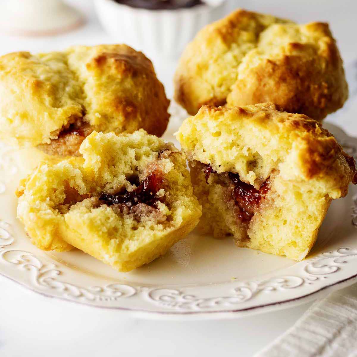 Three biscuit muffins on a white plate with one cut in half exposing the jam inside