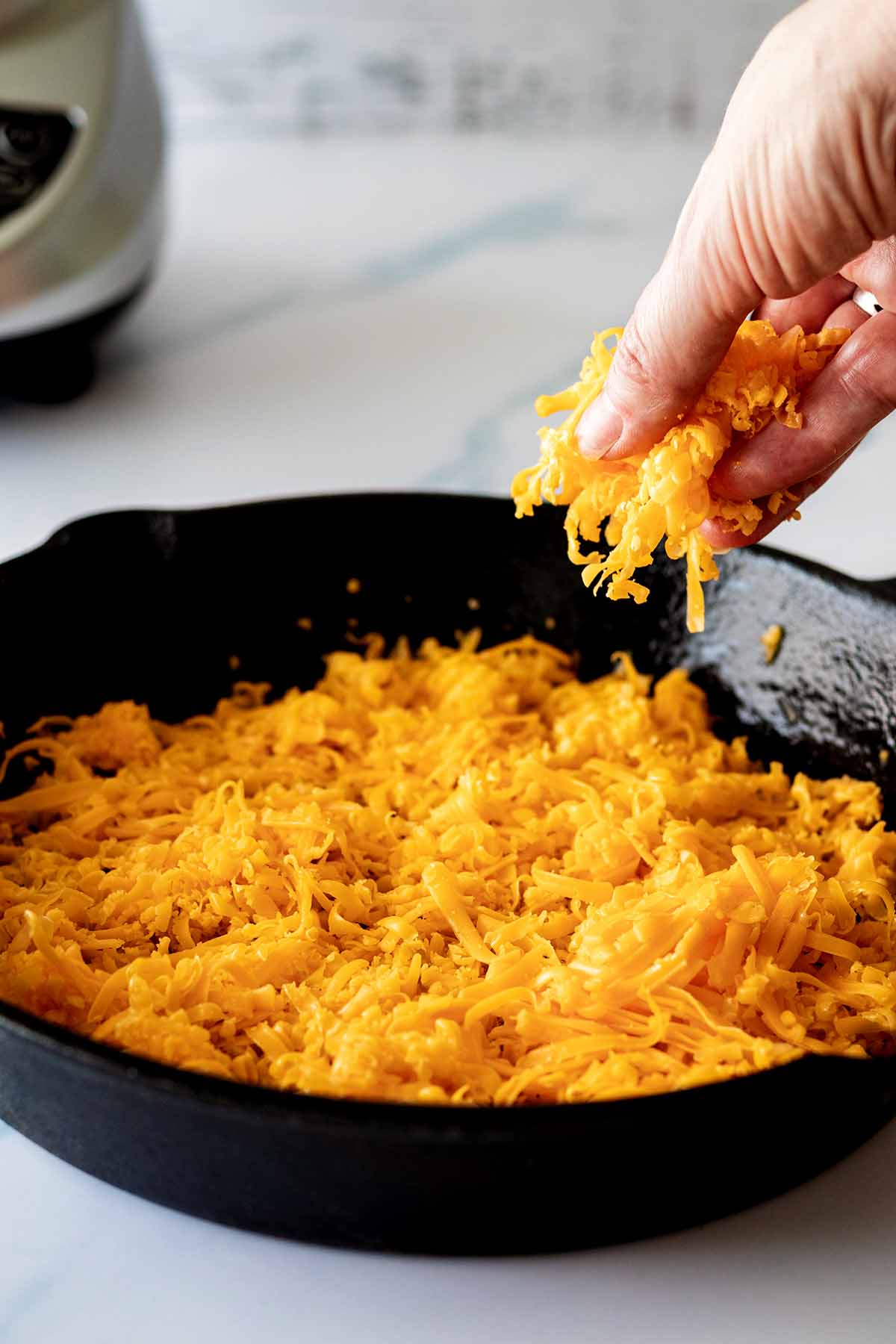 Grated cheddar cheese being sprinkled on the bottom of a cast iron skillet