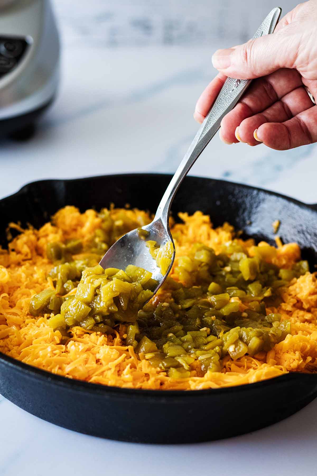 Green chiles being spooned over cheddar cheese in a cast iron skillet