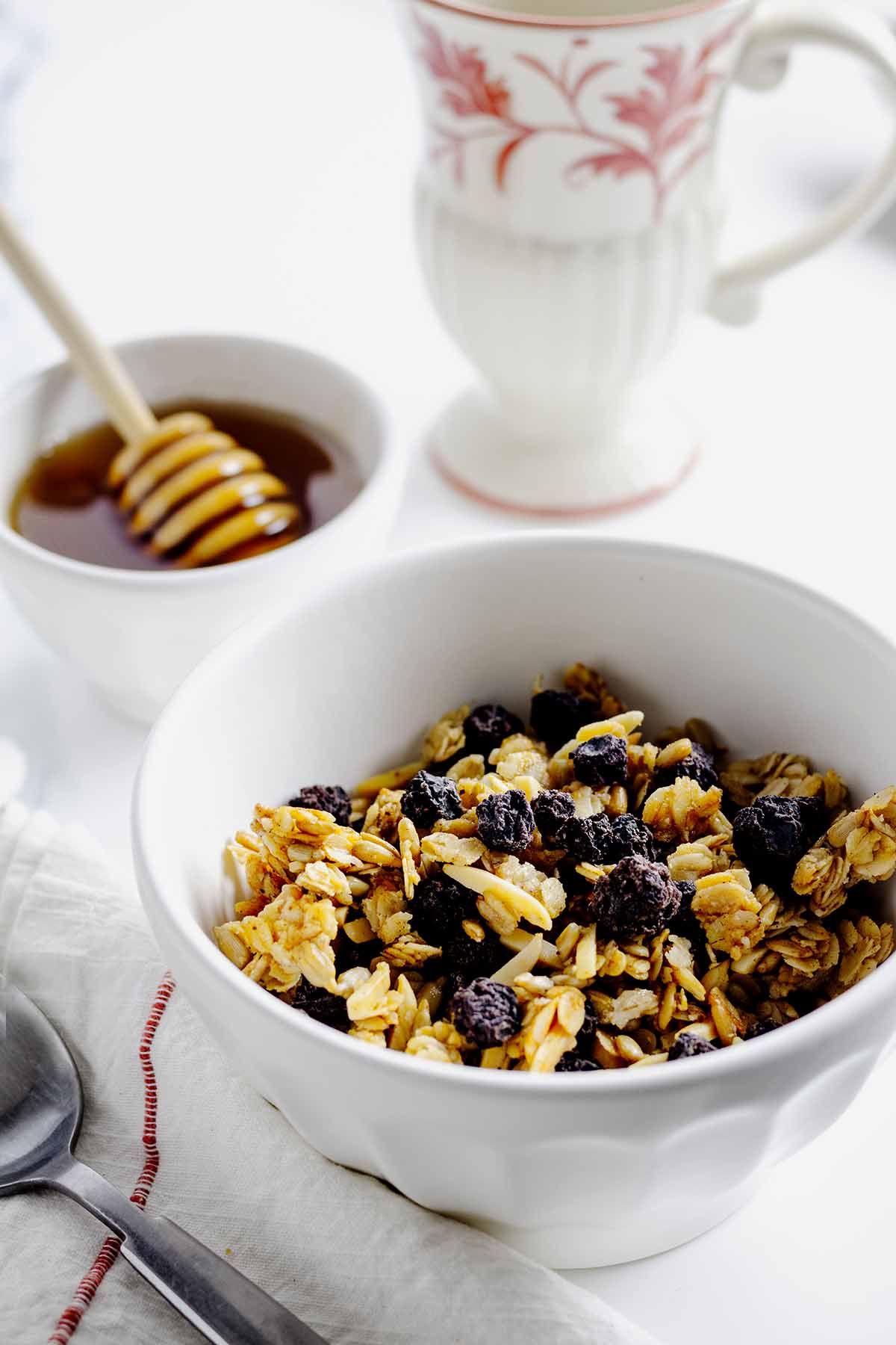 Blueberry granola in a white bowl with a small bowl of honey and wand in the background