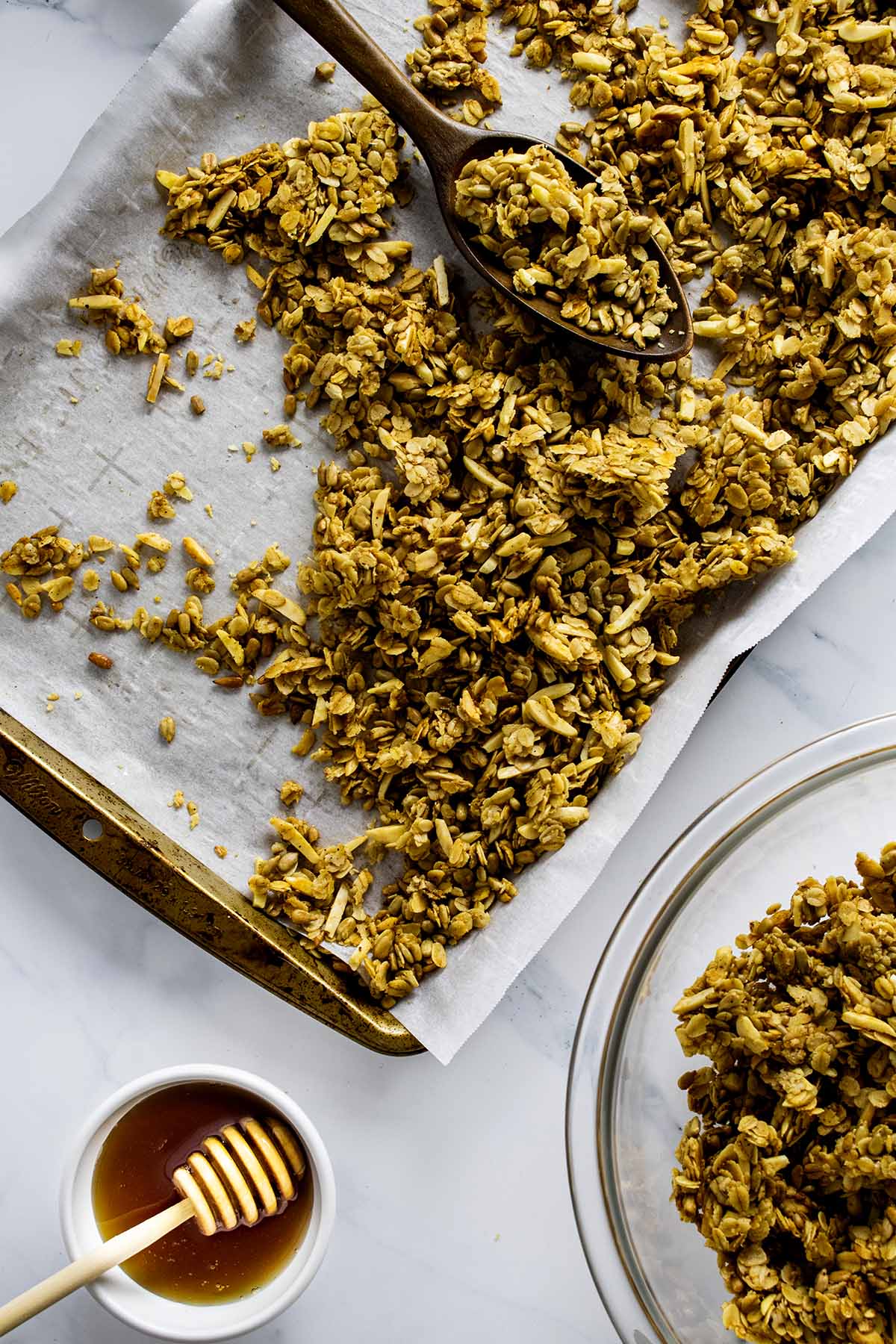 Overhead photo of granola clusters on a baking dish with a bowl of clusters and small bowl of honey with honey wand