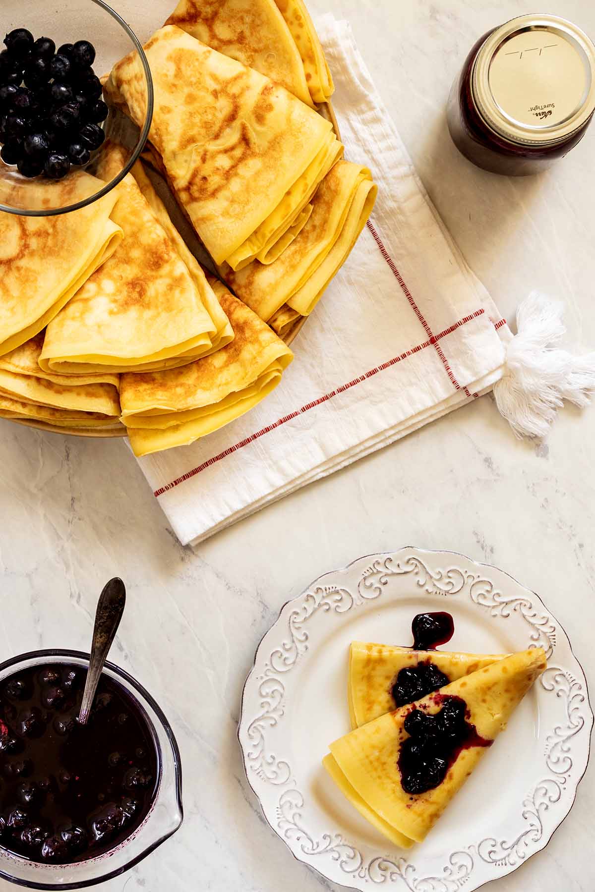 Overhead view of folded breakfast plates on a platter and a single crepe topped with blueberry syrup on a white plate