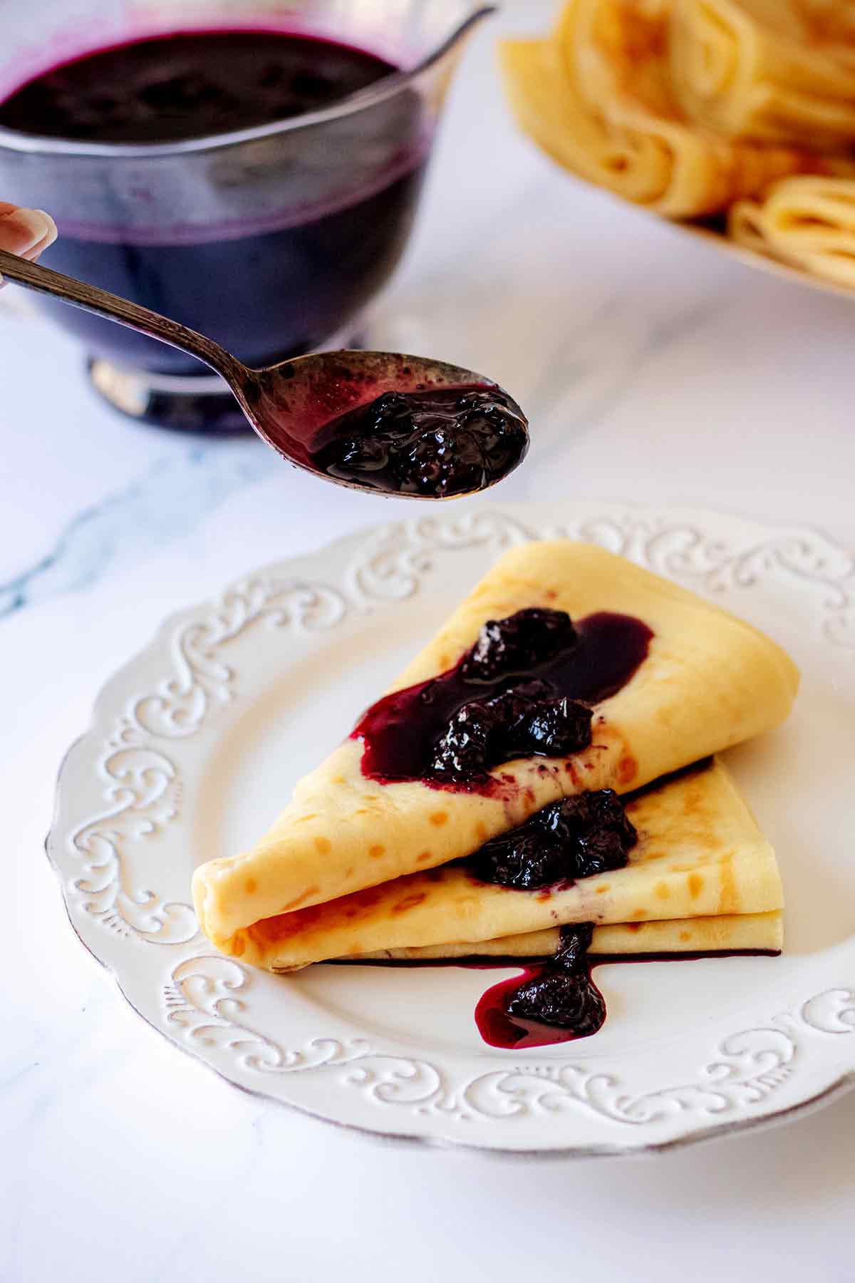 Spoonful of homemade blueberry syrup being poured onto a folded breakfast crepe on a white plate
