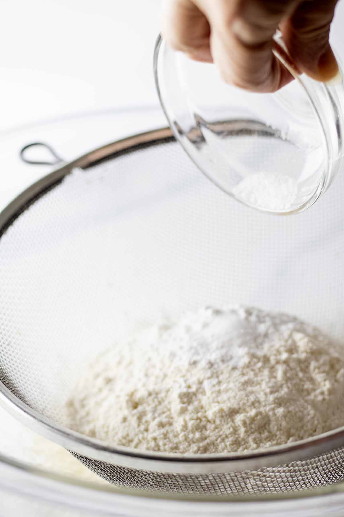 Dry ingredients being sifted through a mesh strainer into a glass bowl
