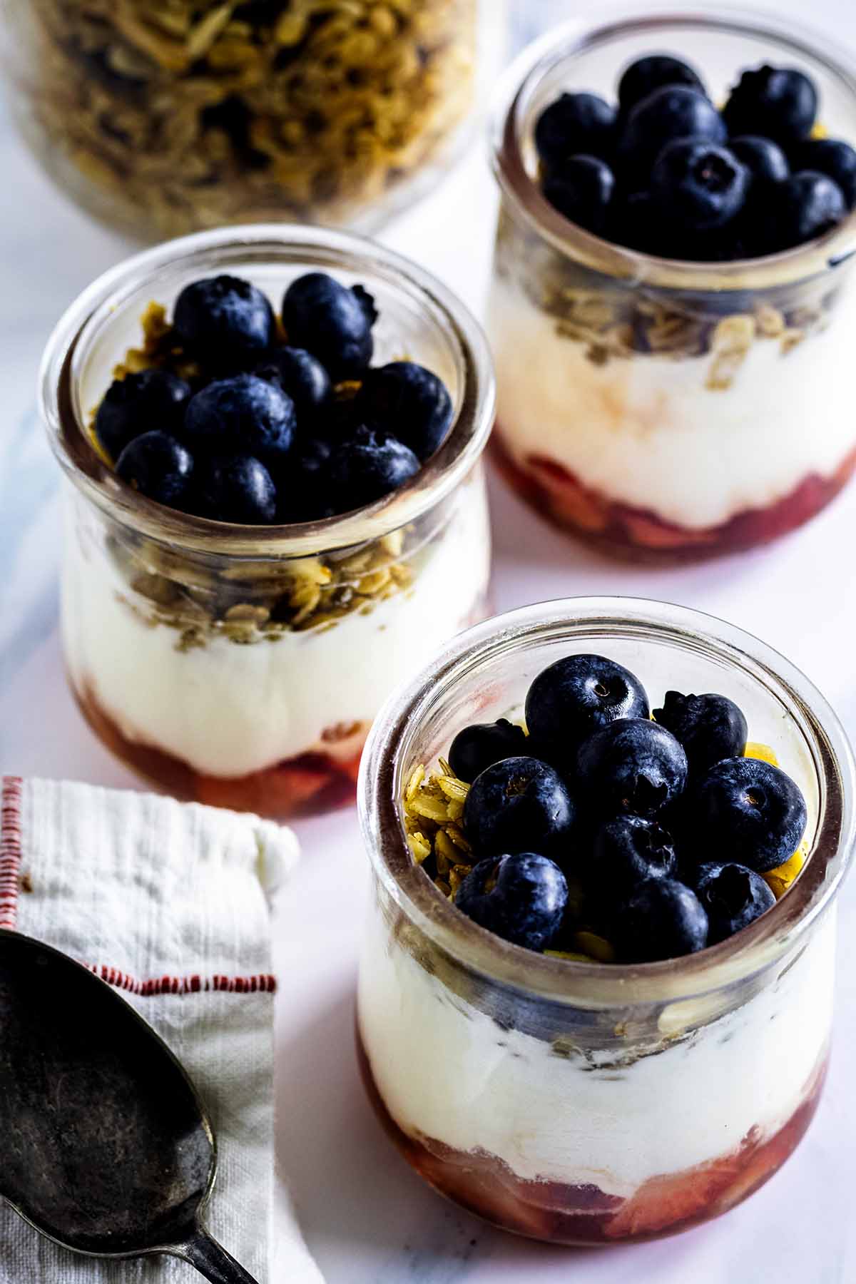 Top view of three finished parfaits in glass jars with a spoon, red and white napkin and granola in a jar