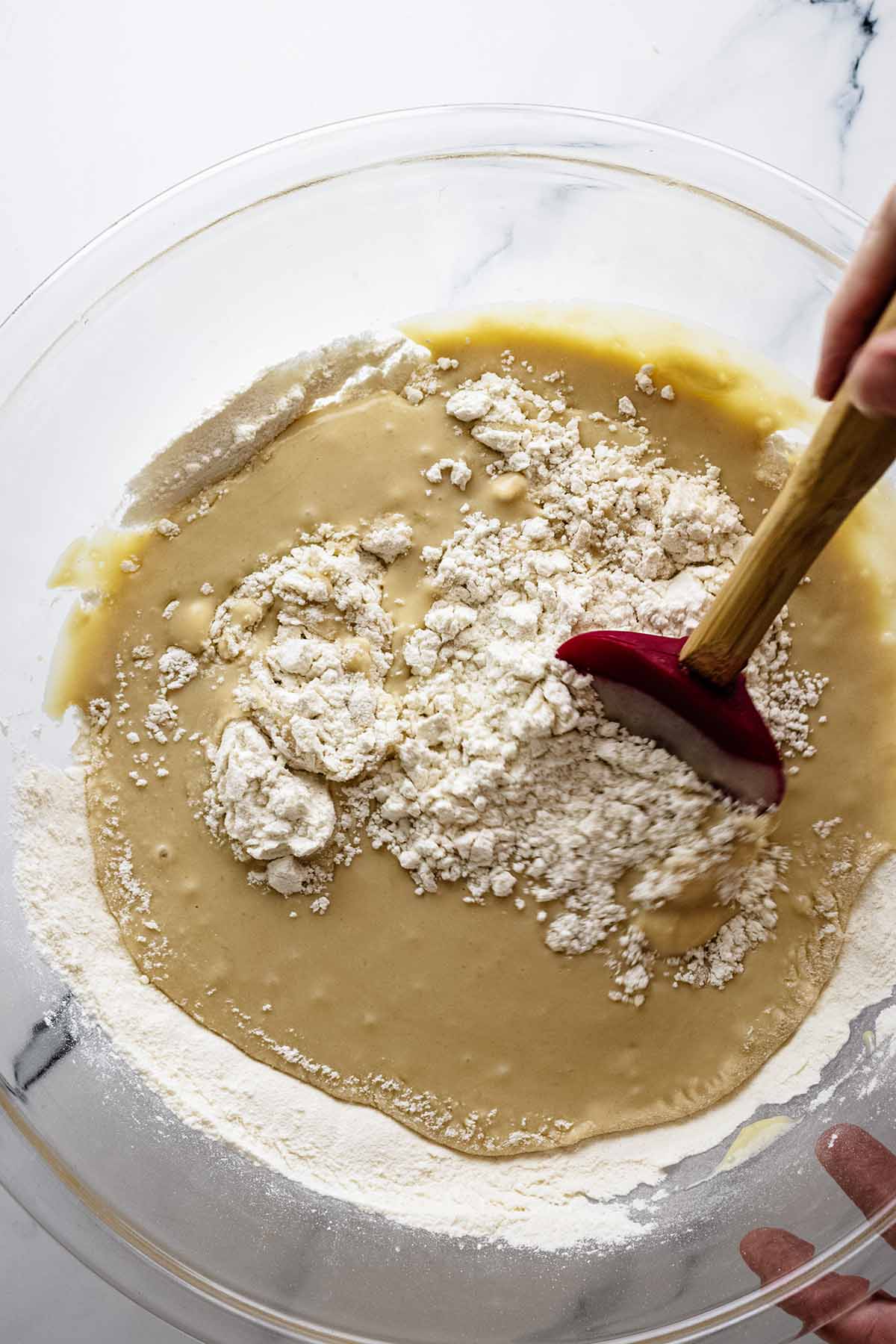 Wet and dry ingredients in a large glass bowl being stirred with a red spatula