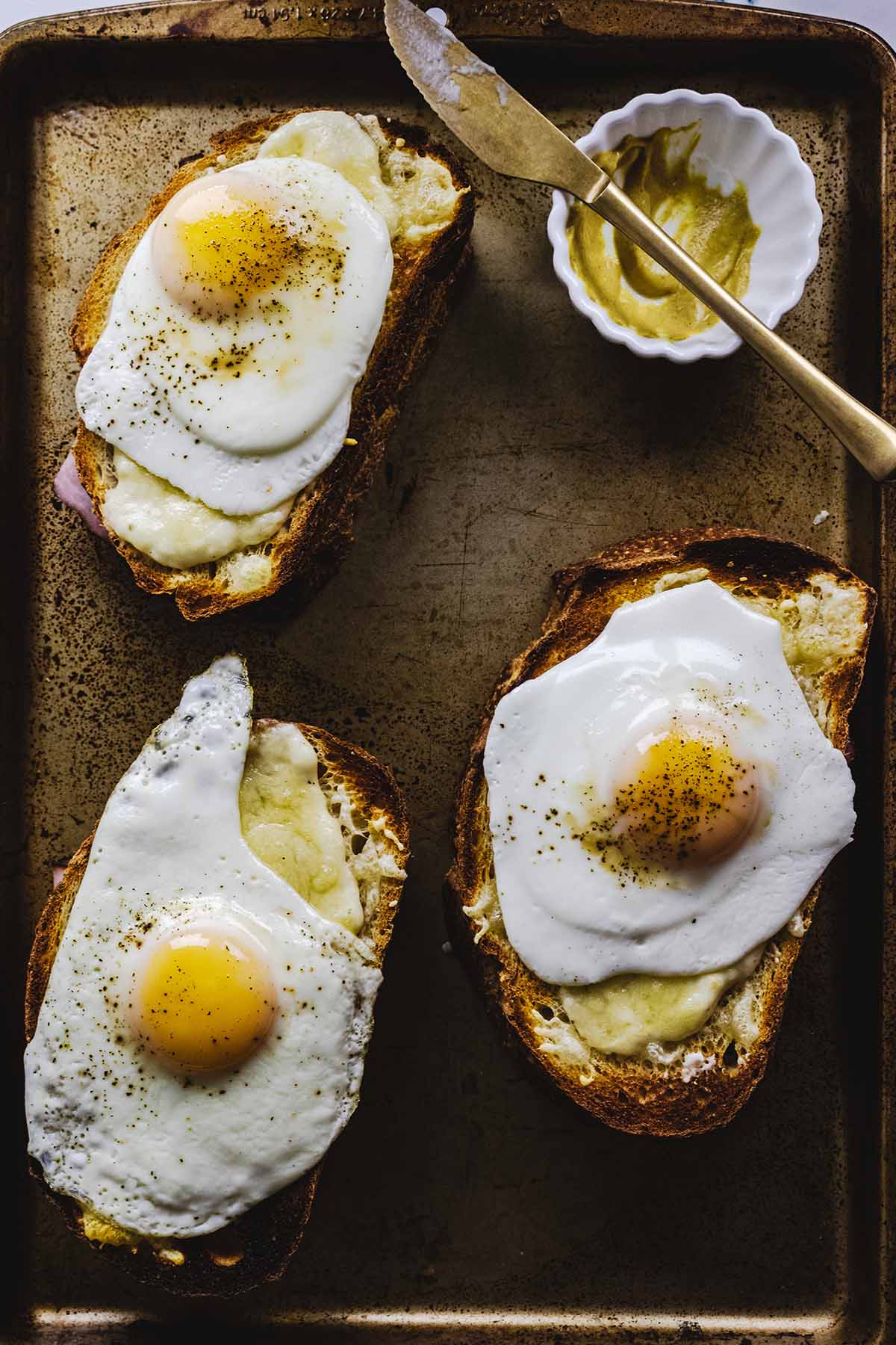 Overhead view of 3 croque madame sandwiches on a cookie sheet with a small bowl of mustard and a gold knife