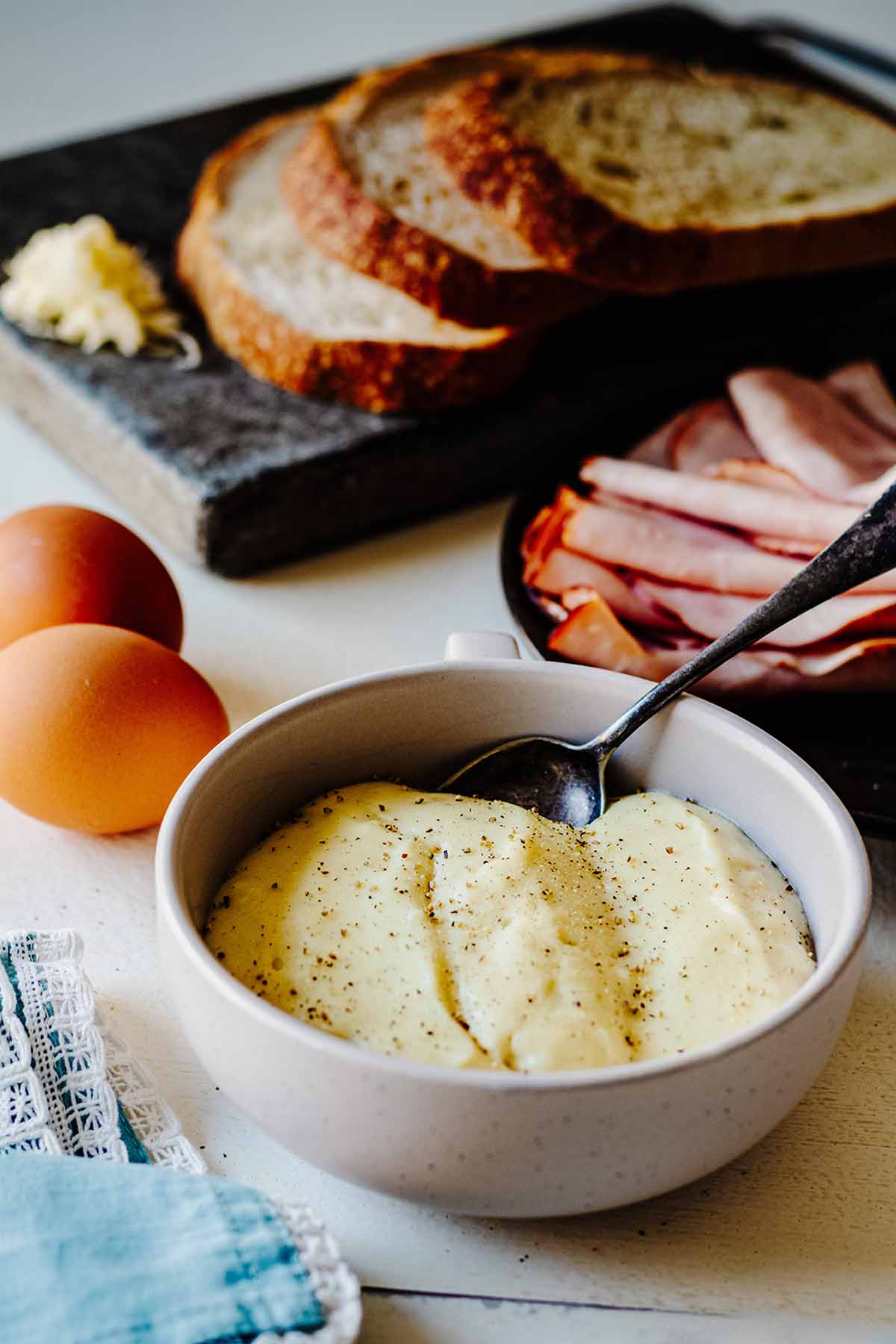 Croque madame sauce in a bowl with a spoon. Ham, eggs and sliced bread are in the background.