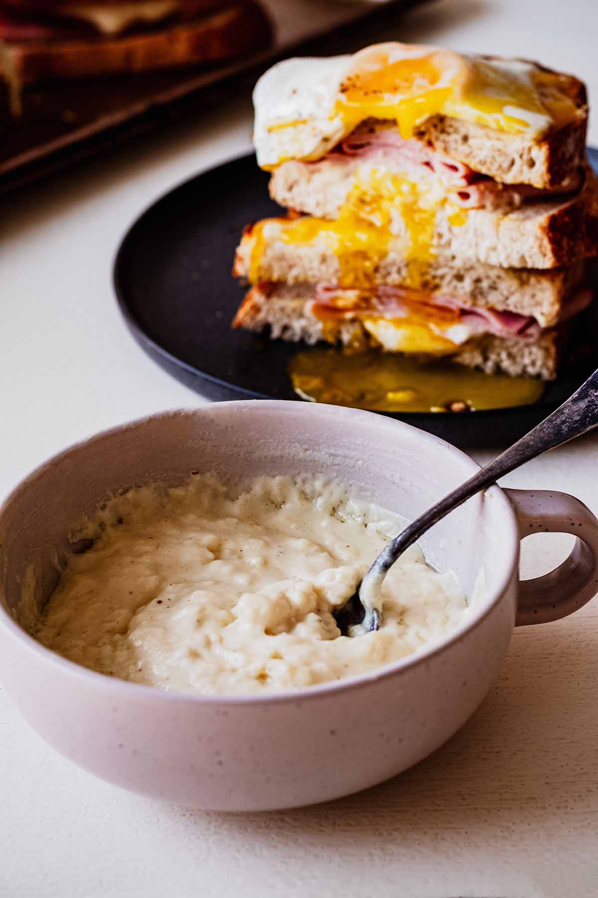 Bowl of croque madame sauce with a spoon. A croque madame sandwich is in the background.