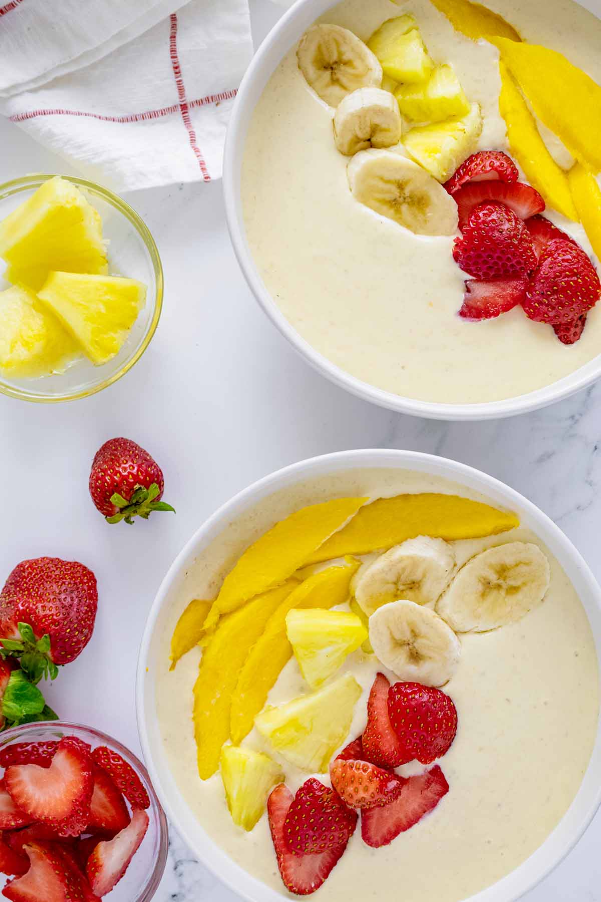 Overhead view of two tropical smoothie bowls in a white bowls with strawberries and pineapple and a white and red napkin.