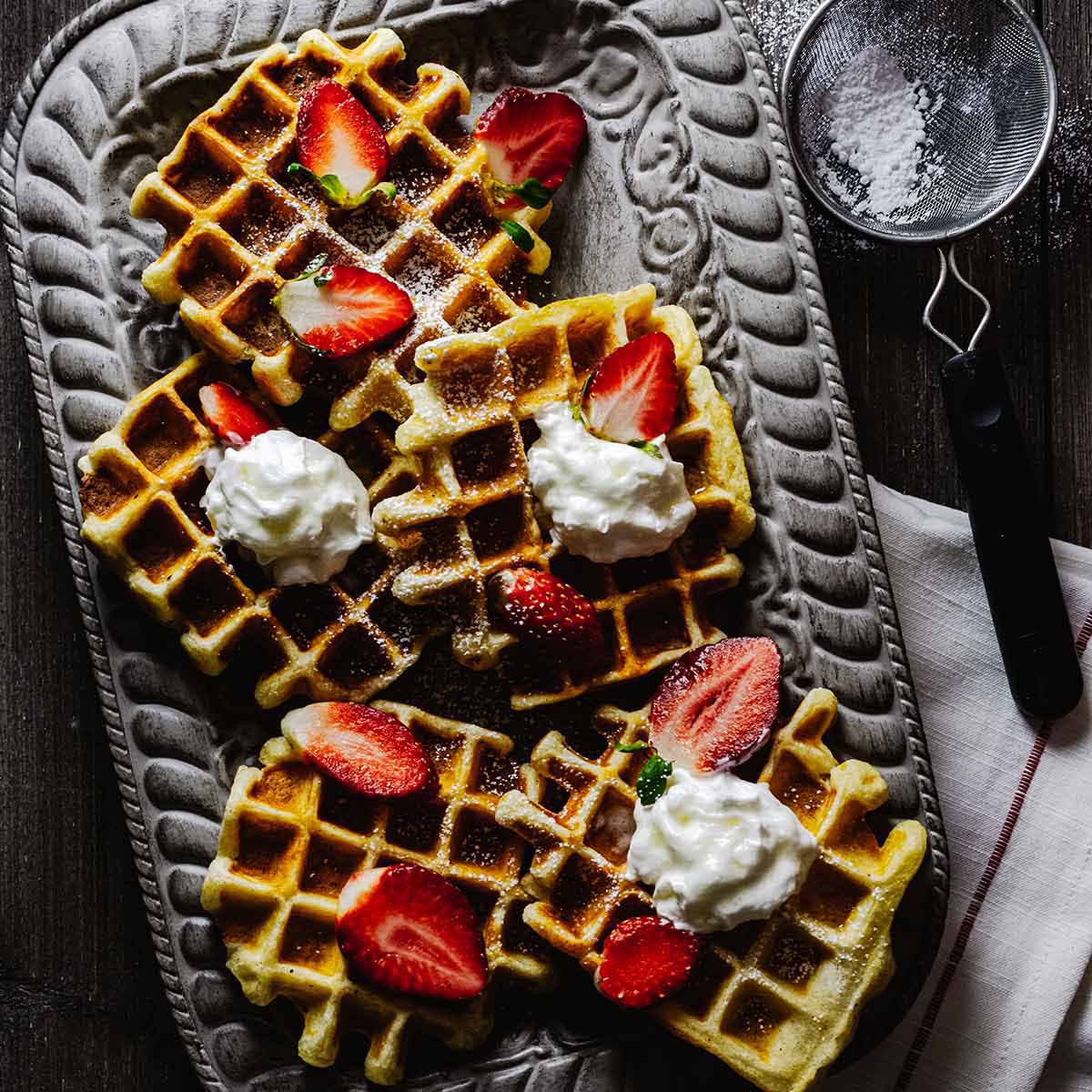 Overhead view of buttermilk Belgian waffles on a gray tray with strawberries and whipped cream