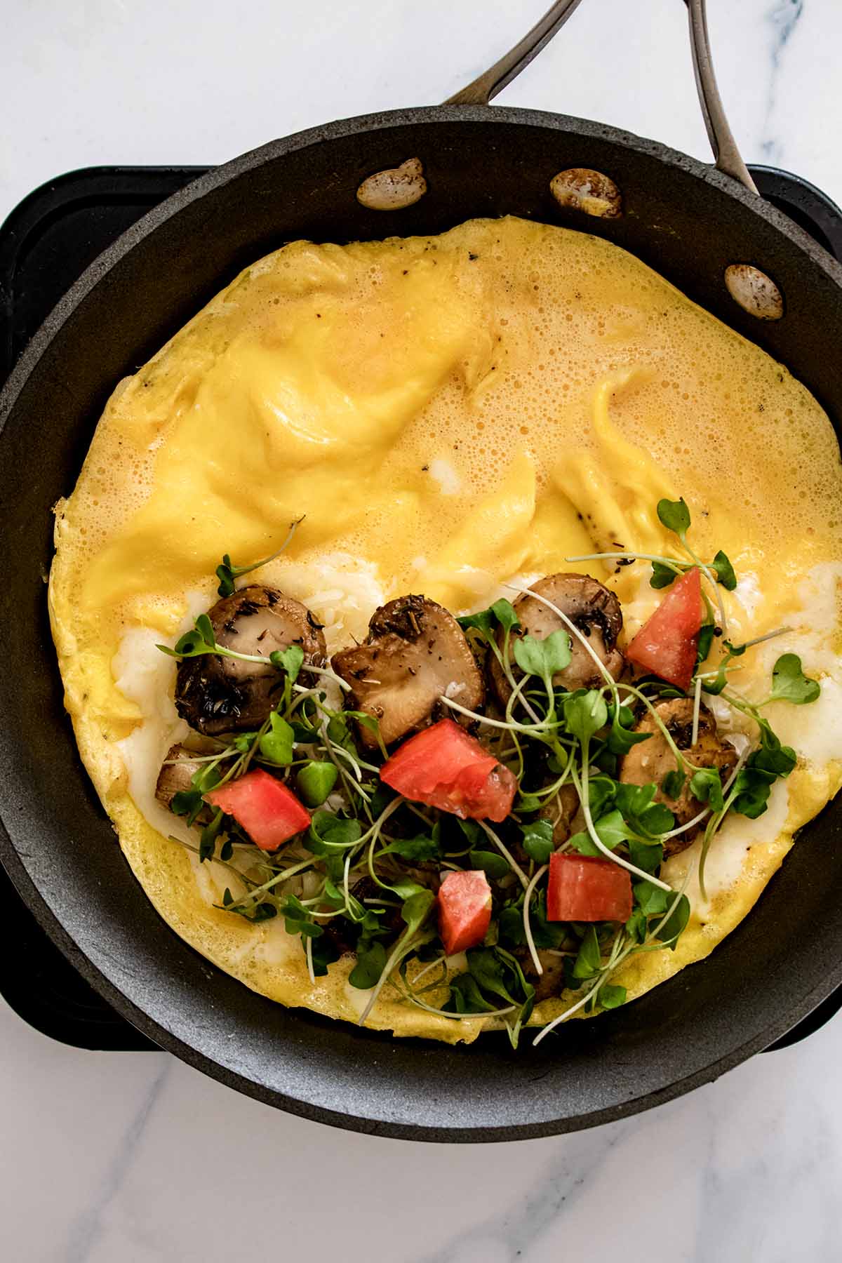 Semi-cooked omelette in a skillet with chopped tomato and micro broccoli added to one half of omelette