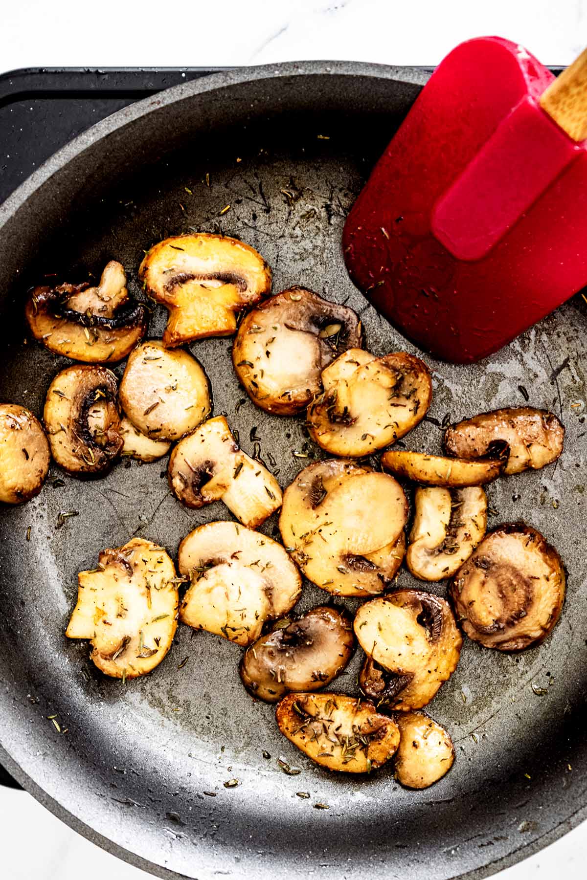 Sliced mushrooms and herbs in a skillet with a red spatula