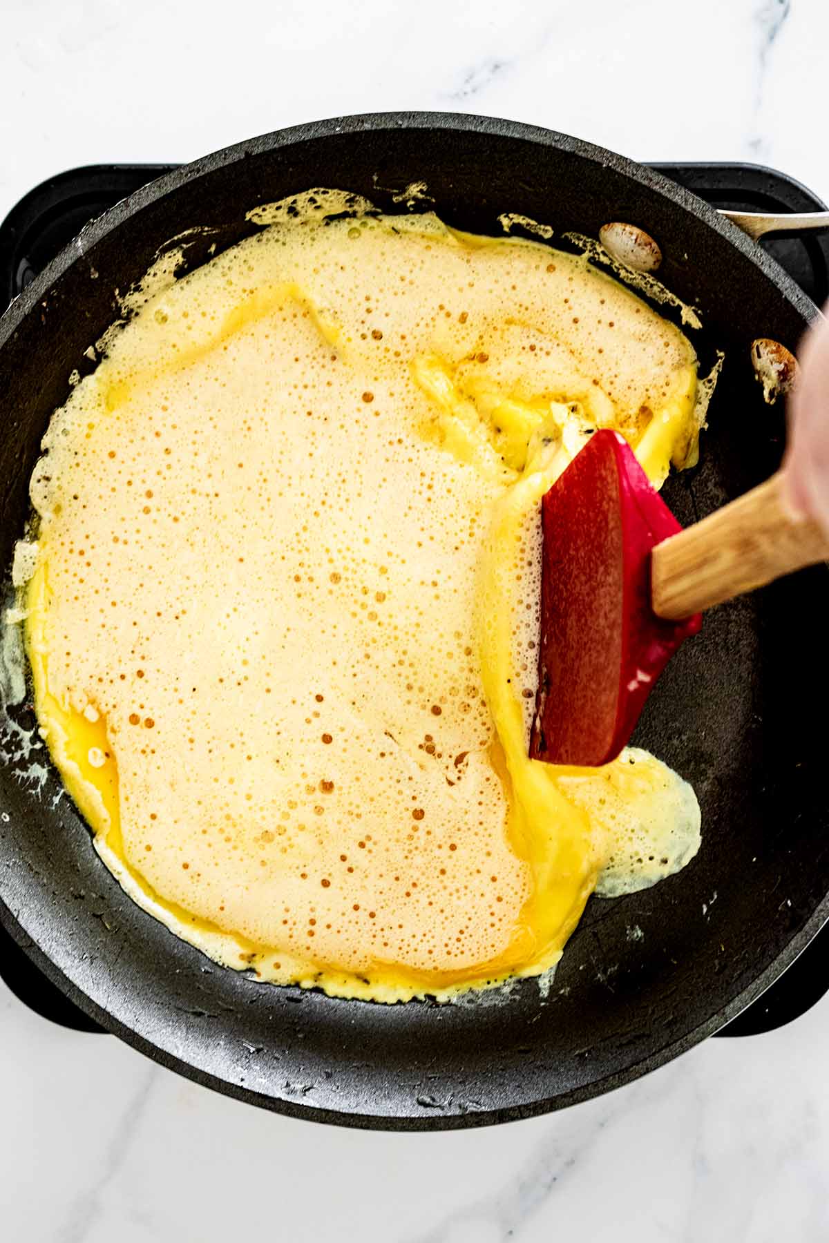 Red spatula pushing edge of semi-cooked eggs toward the center of a skillet