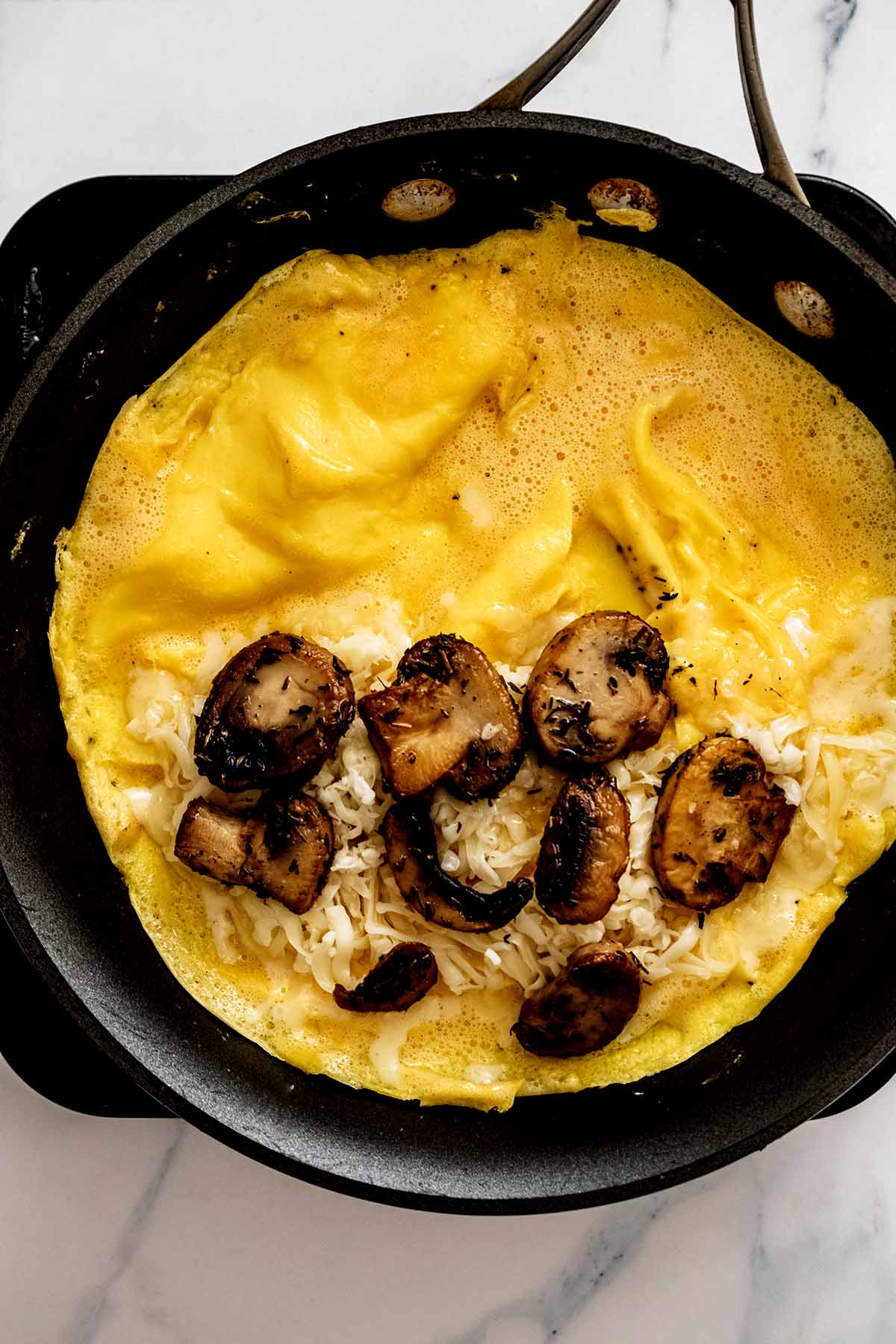 Semi-cooked omelette in a skillet with cooked mushrooms added to one half of omelette