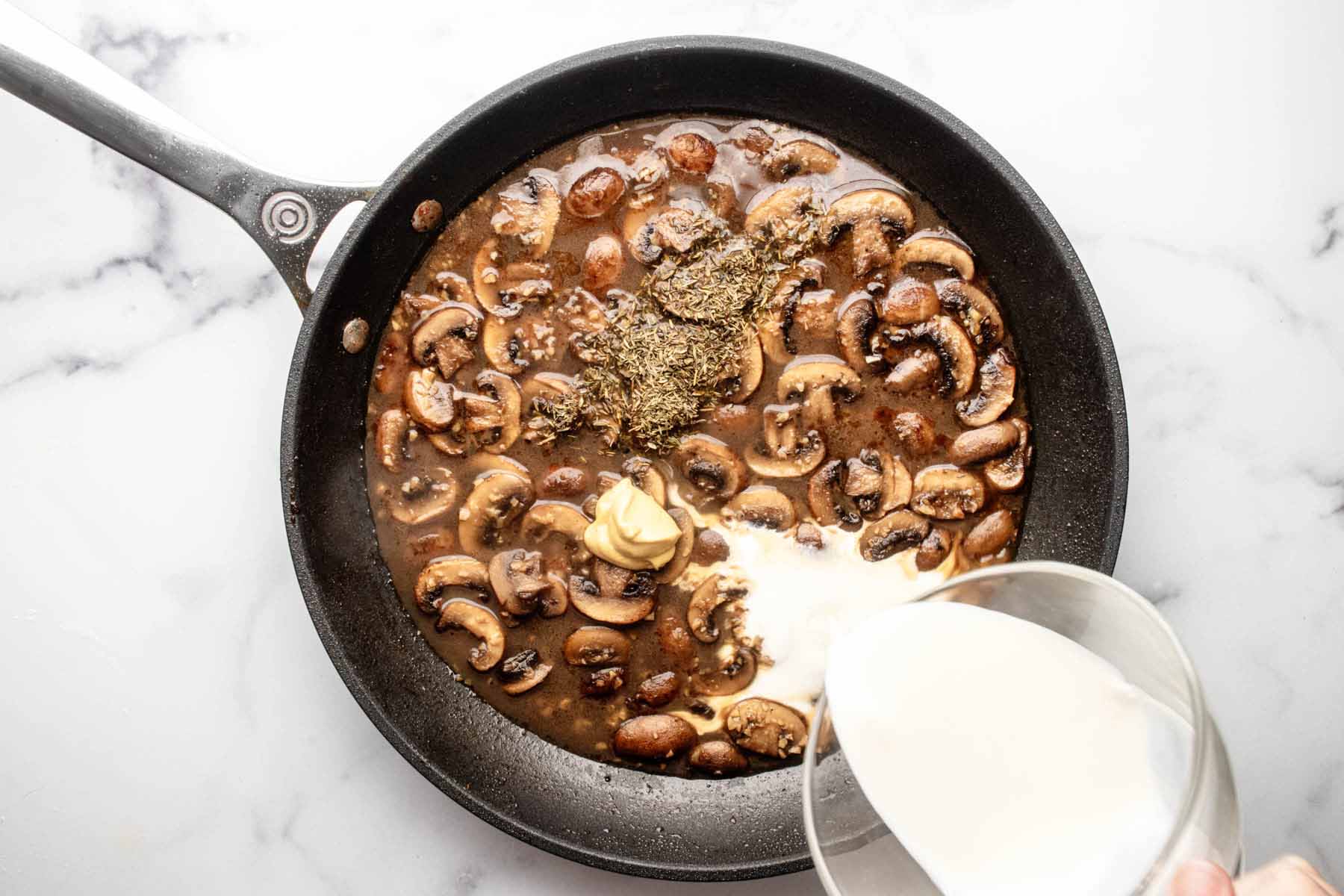 Heavy cream being poured into skillet with mushrooms, mustard, and thyme leaves.