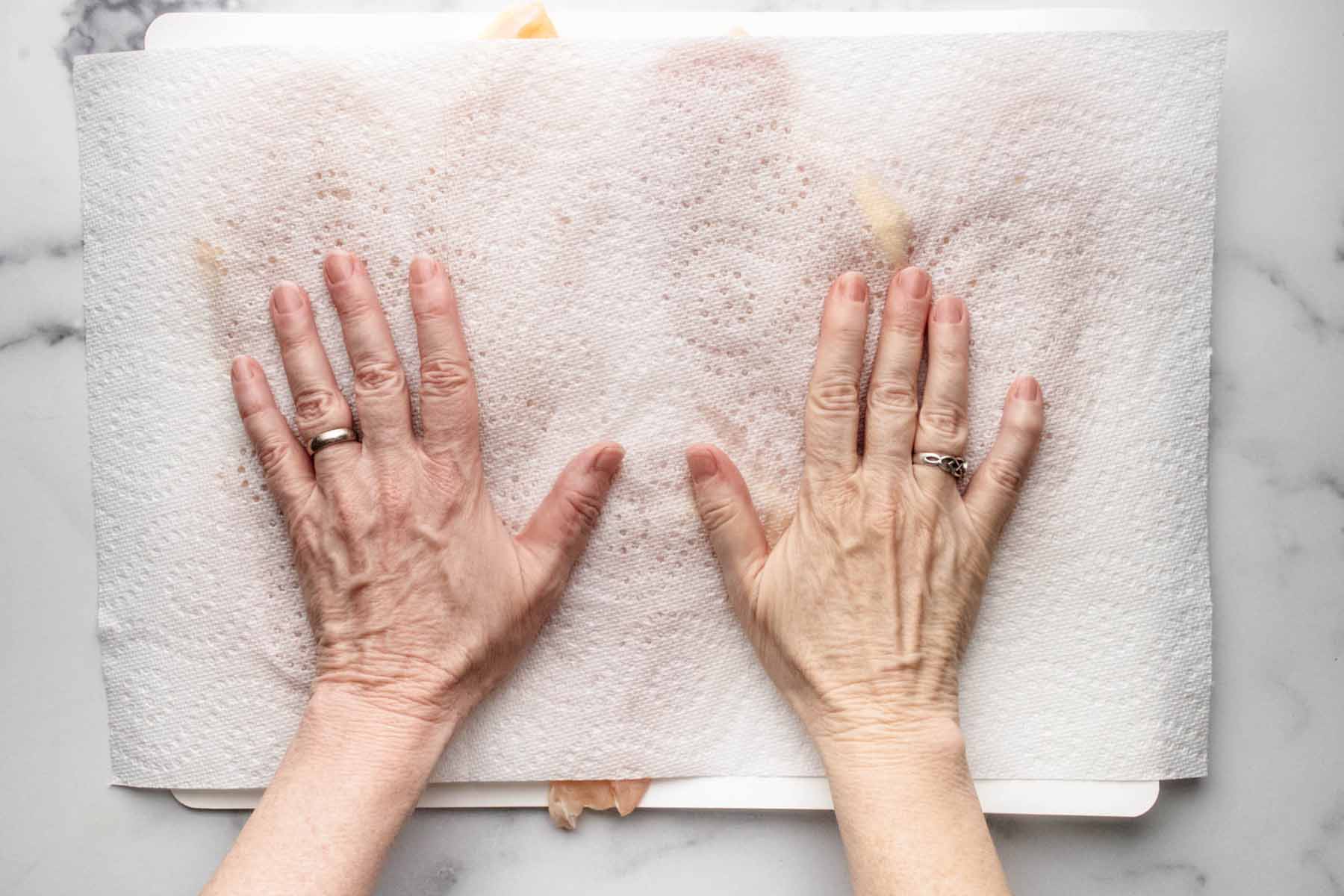 Paper towels on top of chicken cutlets with hands pressing down on the towels.