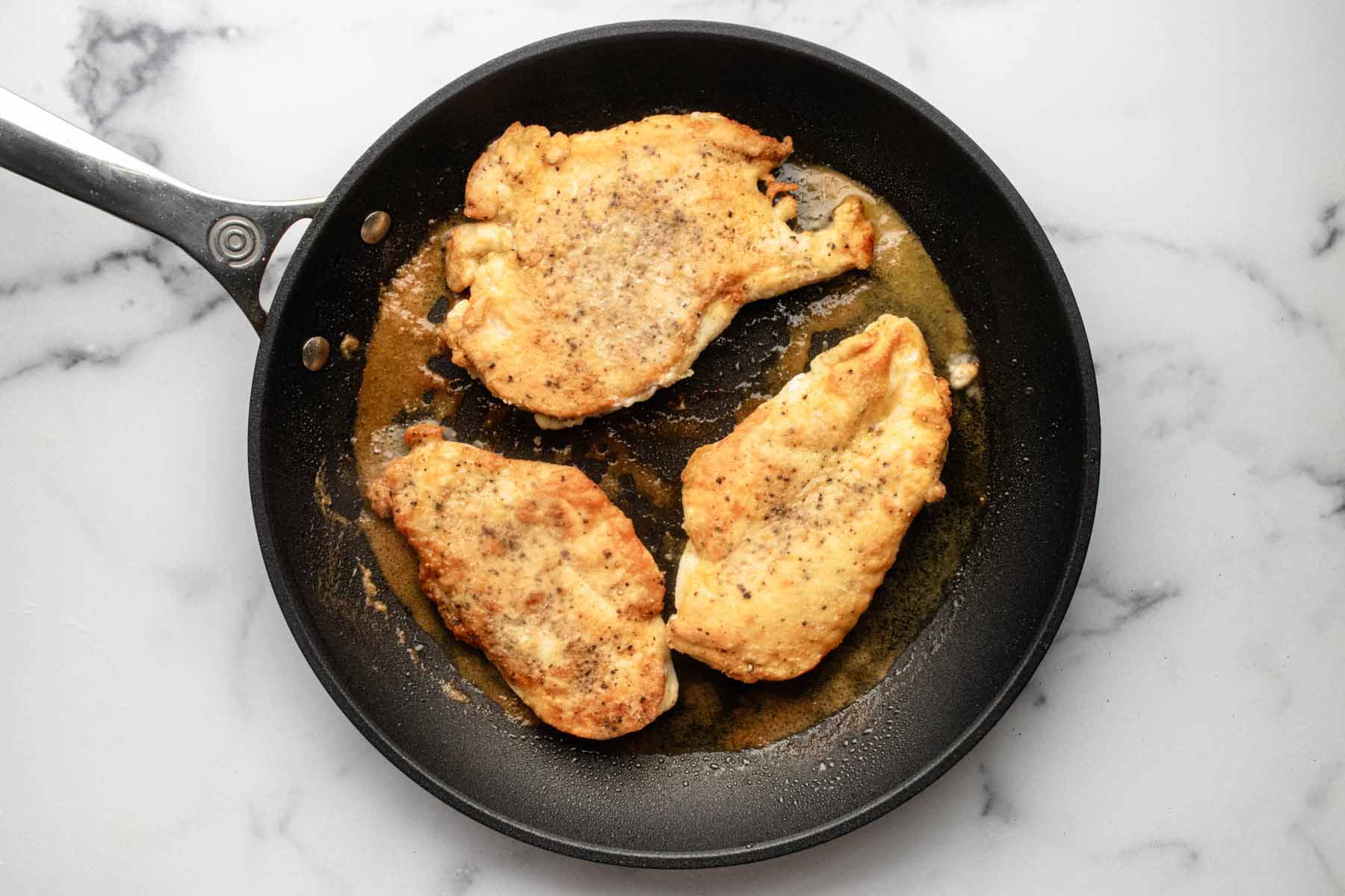 Three browned chicken cutlets cooking in a skillet.