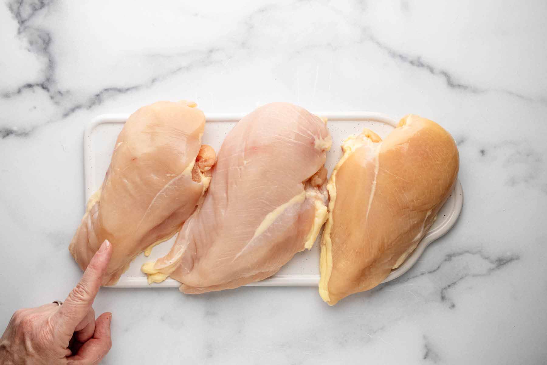 Three chicken breasts on a marble background with a finger pointing to the smallest one.